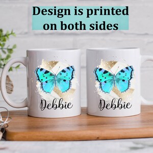 May include: Two white ceramic mugs with a blue butterfly design and gold accents. The mugs have the name "Debbie" printed on them.