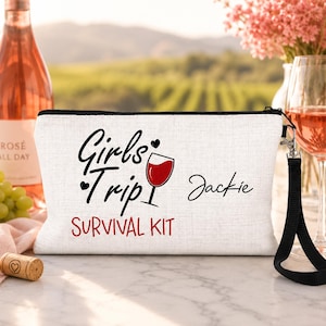 May include: A white rectangular zippered pouch with a black wrist strap. The front features the black text "Girls Trip" with a red wine glass graphic, the name "Jackie" in black script, and the red text "SURVIVAL KIT" printed below.