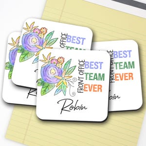 May include: Square white coasters with floral designs and the text "BEST TEAM EVER" in blue, green, and orange. One coaster has the name "Robin" in script. The coasters have a watercolor style with purple and yellow flowers.