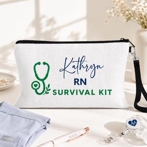 May include: A white rectangular zippered pouch with a black wrist strap. The front features a green stethoscope graphic with a heart, the name Kathryn in blue script, and the text RN SURVIVAL KIT in blue and green block lettering.