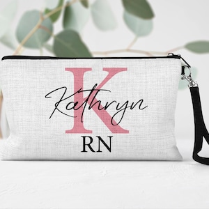 May include: White canvas makeup bag with a pink letter K and the name Kathryn written in black cursive. The bag also has the letters RN in black below the name. The bag has a black zipper and a black strap.