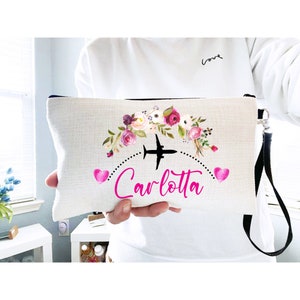 K&ouml;nnte beinhalten: Eine wei&szlig;e Canvas-Rei&szlig;verschlusstasche mit einem schwarzen Rei&szlig;verschluss und einem schwarzen Riemen. Die Tasche hat ein Blumenmuster mit rosa Blumen und einem schwarzen Flugzeug mit einem rosa Herzen auf jeder Seite des Flugzeugs. Der Text "Carlotta" ist in rosa unter dem Flugzeug geschrieben.
