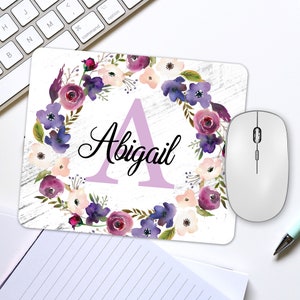 May include: A personalized mousepad featuring the name "Abigail" in black script, with a large lavender letter "A". The design includes a floral wreath in shades of purple, pink, and white, set against a white, distressed wood-look background. A white wireless mouse is next to the mousepad.