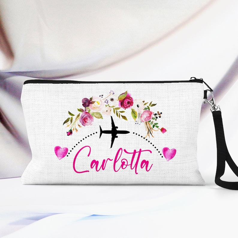 K&ouml;nnte beinhalten: Wei&szlig;e Rei&szlig;verschlusstasche mit schwarzem Rei&szlig;verschluss und schwarzem Trageriemen. Die Tasche hat ein Blumenmuster mit rosa Bl&uuml;ten und einem schwarzen Flugzeug mit einer gepunkteten Linie darum herum. Der Text "Carlotta" ist in rosa unter dem Flugzeug geschrieben.