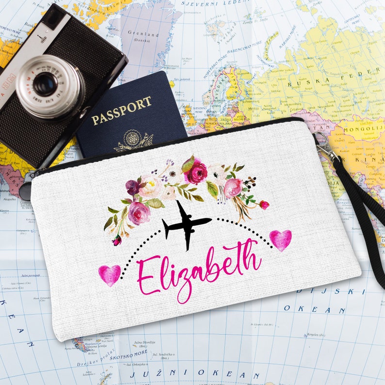 K&ouml;nnte beinhalten: Wei&szlig;e Rei&szlig;verschluss-Reise-Schminktasche mit einem rosafarbenen Blumendesign, einem rosafarbenen Herzen und dem Text "Elizabeth" mit einem Flugzeugsymbol &uuml;ber dem Namen.