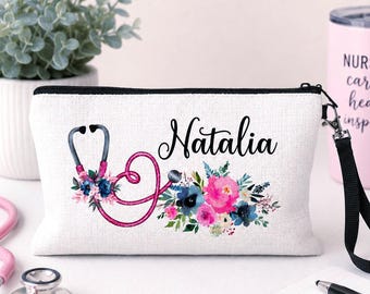 May include: A white rectangular zippered pouch featuring a black script name Natalia, a pink stethoscope graphic, and colorful floral accents in shades of pink and blue. The pouch includes a black wrist strap attached to a silver metal zipper pull.
