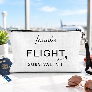 May include: A white linen-textured zippered pouch with a black wrist strap. The front features the black printed text "Laura's FLIGHT SURVIVAL KIT" with a small airplane icon and a dashed flight path line trailing behind the airplane graphic.
