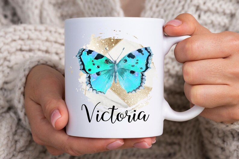 May include: White ceramic mug with a blue butterfly graphic and gold glitter accents. The mug has the name "Victoria" printed on the front.