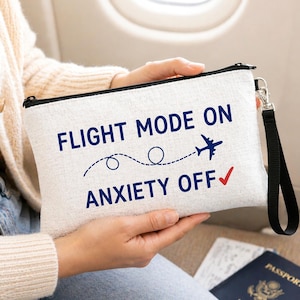 May include: A rectangular off-white canvas pouch with a black zipper and a black wrist strap. The front features dark blue text reading FLIGHT MODE ON and ANXIETY OFF with a red checkmark and a dark blue graphic of an airplane with a dashed flight path.