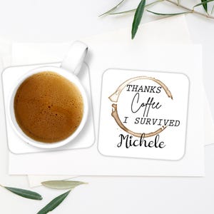 May include: A white ceramic coffee mug filled with coffee sits on a white square coaster. The coaster has a coffee stain design and the text "THANKS Coffee I SURVIVED Michele". A second coaster is next to the mug.
