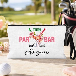 May include: A white canvas zippered pouch with a black wrist strap. The front features a graphic with the text "PAR THEN BAR," a cocktail glass, two golf clubs, and pink flowers. The name "Abigail" is printed in black script below the graphic.