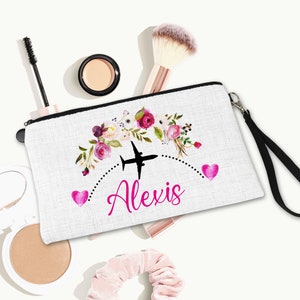 K&ouml;nnte beinhalten: Wei&szlig;e Kosmetiktasche aus Canvas mit schwarzem Rei&szlig;verschluss und schwarzem Trageriemen. Die Tasche hat ein Blumenmuster mit rosa Bl&uuml;ten und ein schwarzes Flugzeug mit dem Namen "Alexis" in rosa.