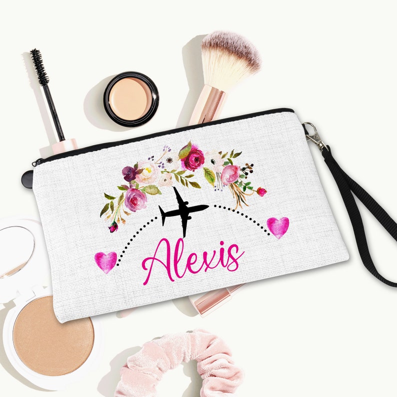 K&ouml;nnte beinhalten: Wei&szlig;e Kosmetiktasche aus Canvas mit schwarzem Rei&szlig;verschluss und schwarzem Trageriemen. Die Tasche hat ein Blumenmuster mit rosa Bl&uuml;ten und ein schwarzes Flugzeug mit dem Namen "Alexis" in rosa.