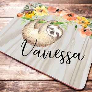 May include: A personalized coaster with a sloth hanging from a branch with watercolor flowers in shades of orange, yellow, and green. The name "Vanessa" is written in black cursive font on a woodgrain background.