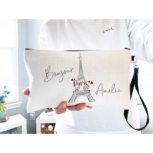 May include: A white canvas clutch with a black wrist strap. The clutch features a black Eiffel Tower illustration with the words "Bonjour Paris Amelie" in black script. Red hearts accent the word "Paris".
