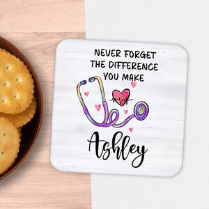 May include: White square coaster with a stethoscope design, a heart with a heartbeat, and the text "Never Forget The Difference You Make" and the name "Ashley". The stethoscope is purple and yellow.