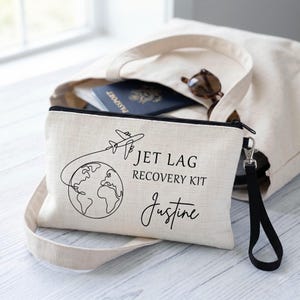 May include: A beige canvas travel pouch with a black zip and wrist strap. The pouch has a black line drawing of an aeroplane circling the globe, with the text "JET LAG RECOVERY KIT" and the name "Justine". A beige tote bag, passport, and sunglasses are in the background.