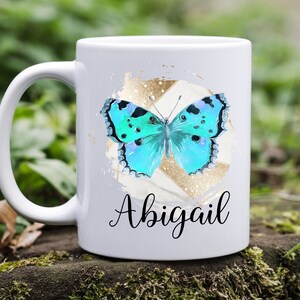 May include: White ceramic mug with a blue butterfly graphic and gold glitter accents. The mug has the name "Abigail" written in black cursive.