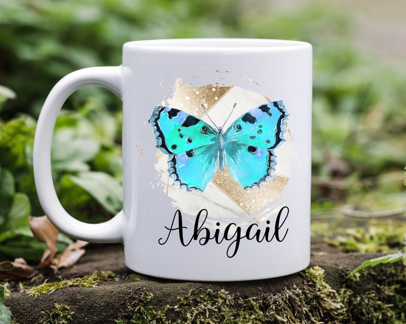 May include: White ceramic mug with a blue butterfly graphic and gold glitter accents. The mug has the name "Abigail" written in black cursive.