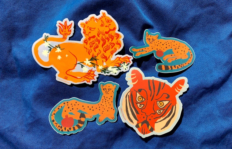 May include: Four colorful stickers featuring a lion, a tiger, and two cheetahs. The lion is orange and has a mane of fire. The tiger has orange and black stripes and three eyes. The cheetahs are orange with black spots. All of the stickers have a white background.