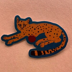May include: A sticker of a brown and orange cheetah with black spots, lying down on a blue background. The cheetah has a red pomegranate on its side.