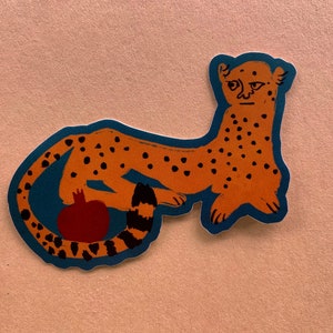 May include: A sticker of a cheetah with a red pomegranate in its tail. The cheetah is orange with black spots and has a blue outline.