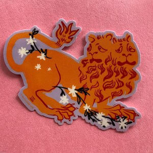 May include: A sticker of a two-headed orange lion with a purple background and white flowers. The lion has a mane of orange flames.