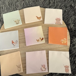 Peut inclure: Plusieurs blocs-notes carrés avec des illustrations d'animaux de la ferme, dont un coq, des poulets et des vaches des Highlands. Les blocs-notes sont de couleurs pastel et disposés sur une surface en bois.