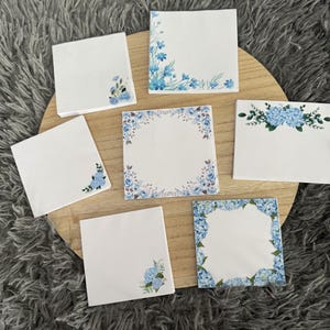 Peut inclure: Bloc-notes carrés blancs avec des motifs floraux bleus. Certains blocs-notes présentent des motifs d'angle, d'autres des bordures ou des arrangements floraux centrés en haut. Les blocs-notes sont disposés sur une surface en bois, sur un fond gris texturé.