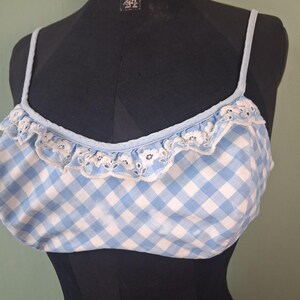 May include: A light blue and white gingham crop top with thin straps and a ruffled edge. The top has a white lace trim along the neckline. The garment is displayed on a black mannequin.