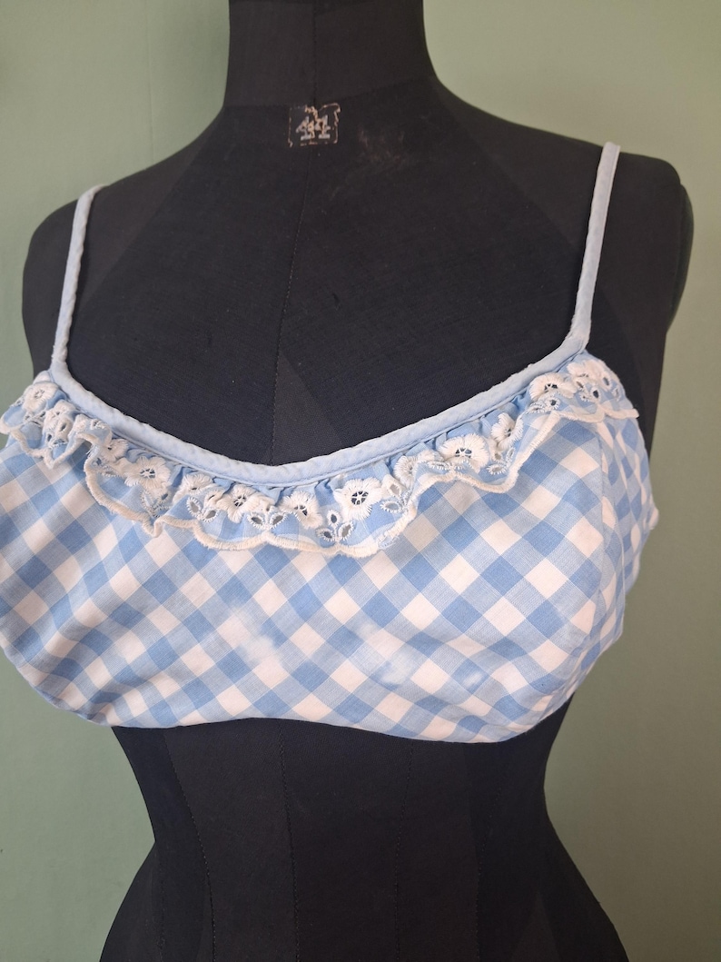May include: A light blue and white gingham crop top with thin straps and a ruffled edge. The top has a white lace trim along the neckline. The garment is displayed on a black mannequin.