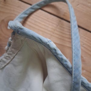 May include: Close-up of a white denim top with a light blue strap and trim. The strap is stitched to the top, and the fabric appears to be soft and worn. The background is a wooden surface.