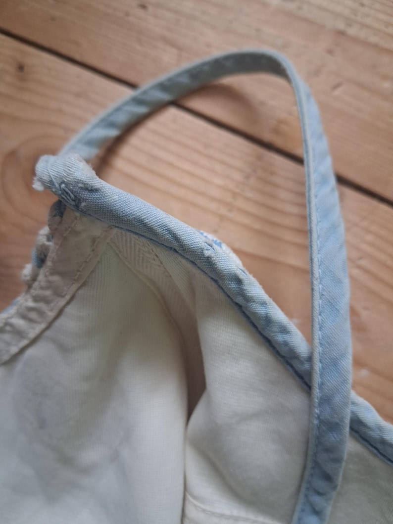 May include: Close-up of a white denim top with a light blue strap and trim. The strap is stitched to the top, and the fabric appears to be soft and worn. The background is a wooden surface.