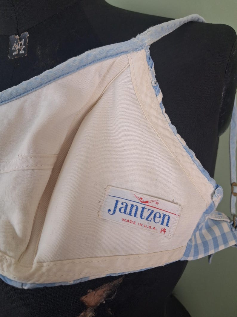 May include: Vintage Jantzen bra in cream and light blue. The bra features a classic design with a light blue trim and straps. The label reads "Jantzen MADE IN U.S.A. 14". The bra is displayed on a mannequin.