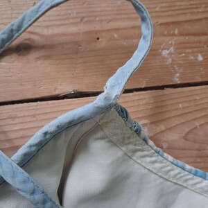 May include: Close-up of a light blue denim top with white lining. The top features thin, adjustable straps and a metal ring detail. The fabric appears worn, with visible stitching and a faded appearance, suggesting a vintage style.