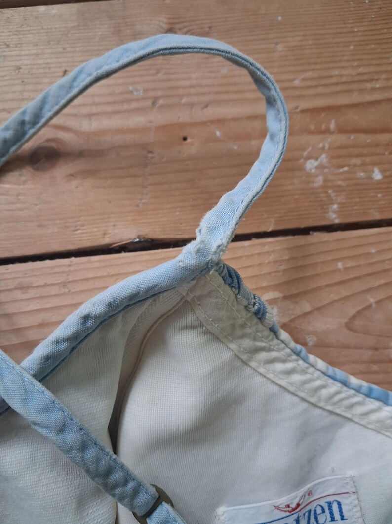 May include: Close-up of a light blue denim top with white lining. The top features thin, adjustable straps and a metal ring detail. The fabric appears worn, with visible stitching and a faded appearance, suggesting a vintage style.