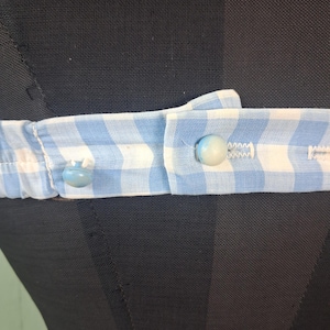 May include: Close-up of a light blue and white gingham fabric belt with two light blue buttons. The belt is fastened and appears to be part of a garment. The background is a black mannequin.