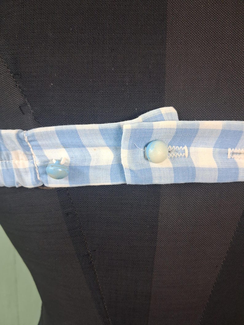 May include: Close-up of a light blue and white gingham fabric belt with two light blue buttons. The belt is fastened and appears to be part of a garment. The background is a black mannequin.