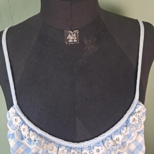 May include: A light blue and white gingham patterned top with delicate white floral lace trim. The top has thin, light blue straps and is displayed on a black mannequin. The overall style is vintage and feminine.