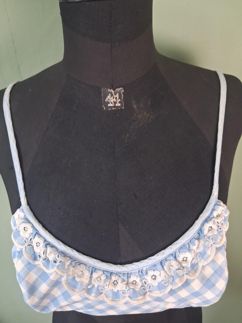 May include: A light blue and white gingham patterned top with delicate white floral lace trim. The top has thin, light blue straps and is displayed on a black mannequin. The overall style is vintage and feminine.
