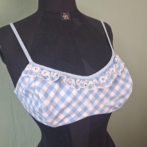 May include: A light blue and white gingham pattern crop top with thin straps and a ruffled trim. The top is displayed on a black mannequin. The trim has a delicate, eyelet design.