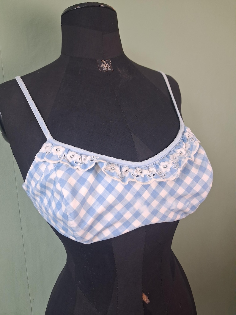 May include: A light blue and white gingham pattern crop top with thin straps and a ruffled trim. The top is displayed on a black mannequin. The trim has a delicate, eyelet design.