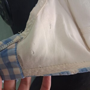 May include: Close-up of a vintage bra. The bra features a white interior and a light blue and white gingham pattern on the exterior. The edges are trimmed with a white fabric.