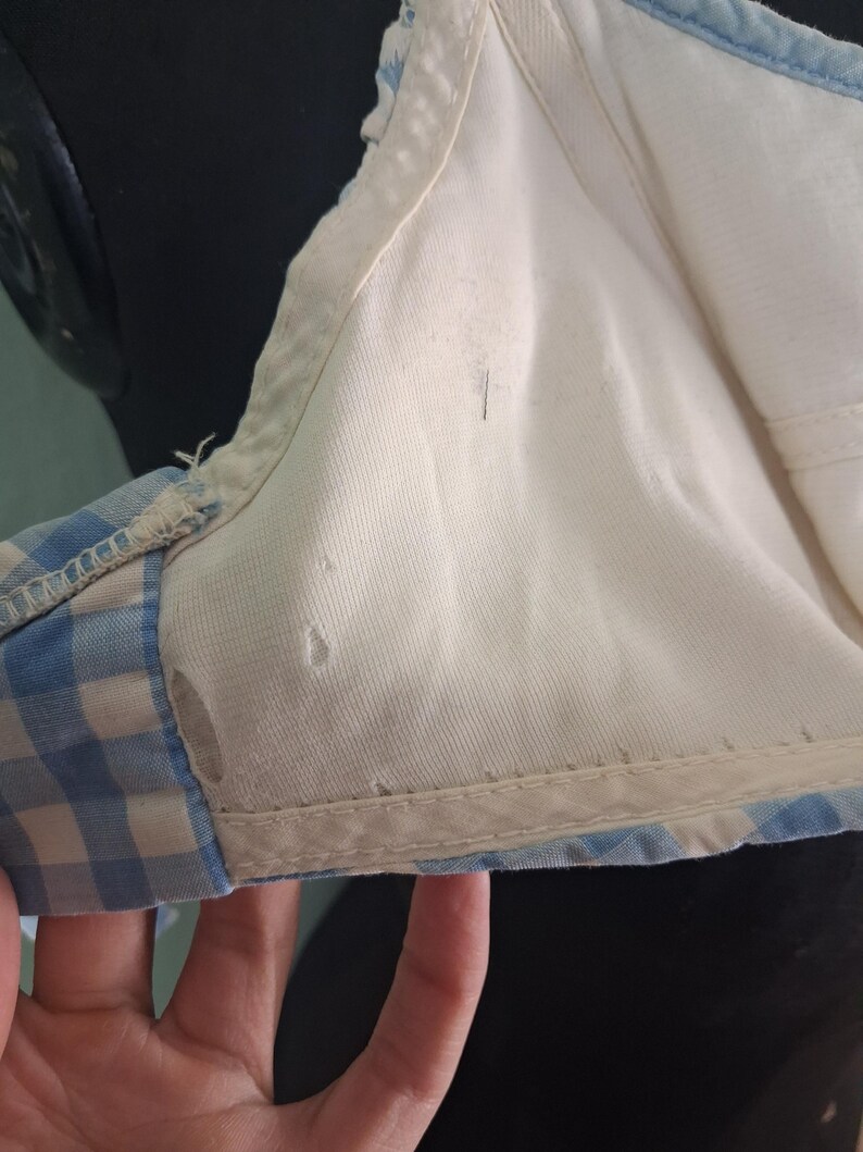 May include: Close-up of a vintage bra. The bra features a white interior and a light blue and white gingham pattern on the exterior. The edges are trimmed with a white fabric.
