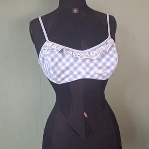 May include: A light blue and white gingham pattern bralette with thin straps and a ruffled trim. The bralette is displayed on a black mannequin against a muted green background. The top has a delicate, feminine design.