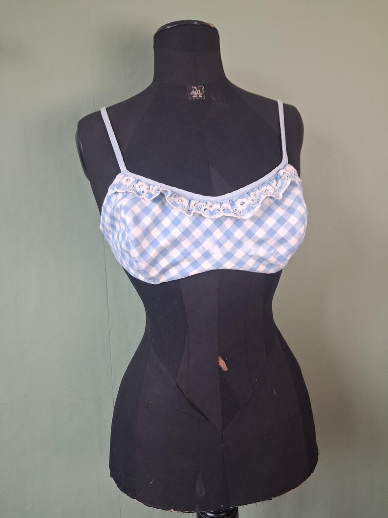 May include: A light blue and white gingham pattern bralette with thin straps and a ruffled trim. The bralette is displayed on a black mannequin against a muted green background. The top has a delicate, feminine design.