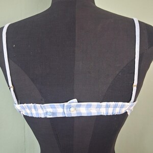 May include: A blue and white gingham pattern bra with adjustable straps. The bra is displayed on a black mannequin. The straps are white and the bra has a button closure in the back.