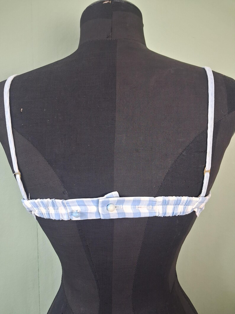 May include: A blue and white gingham pattern bra with adjustable straps. The bra is displayed on a black mannequin. The straps are white and the bra has a button closure in the back.