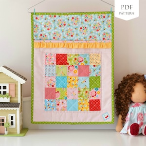 May include: A pink and green patchwork quilt with a floral pattern. The quilt is hanging on a wall with a doll and a toy house in the foreground.