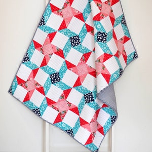 Puede incluir: Una colcha de retazos blanca, roja, rosa y azul con un patrón de estrellas. La colcha está doblada y drapeada sobre una escalera blanca.